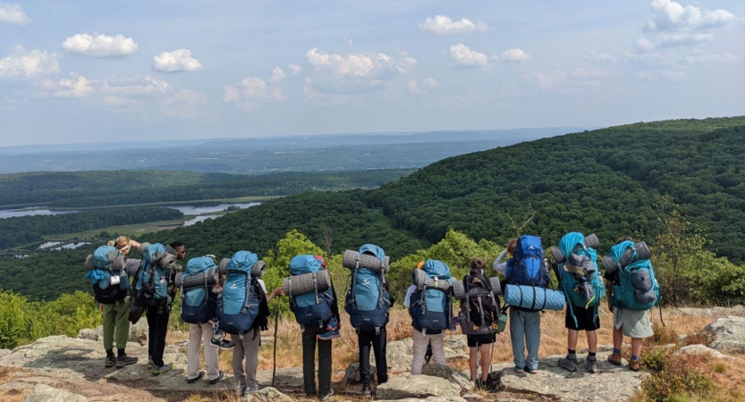 A group of people wearing backpacks stand in a line on a rocky overlook. Below them is vast green landscape, with a river winding through it. 